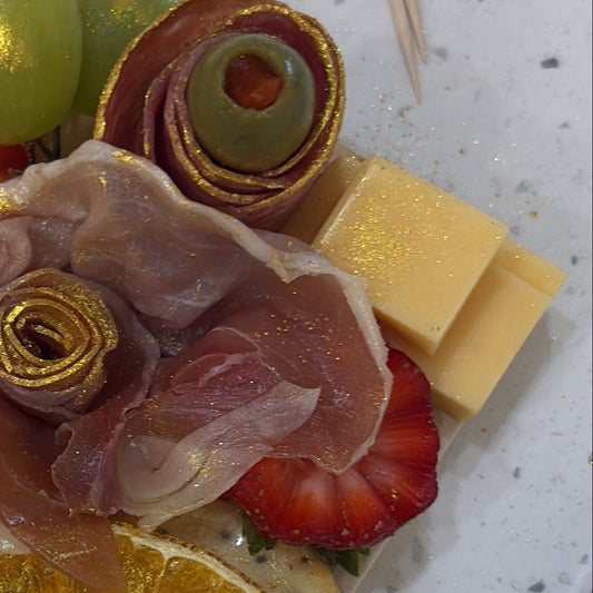 A gourmet charcuterie board or cheese platter arranged on a speckled white countertop, featuring sliced orange, green grapes, strawberries, cheese cubes, rolled prosciutto, and a rolled olive with gold leaf detailing. A toothpick and part of a phone are visible in the frame.